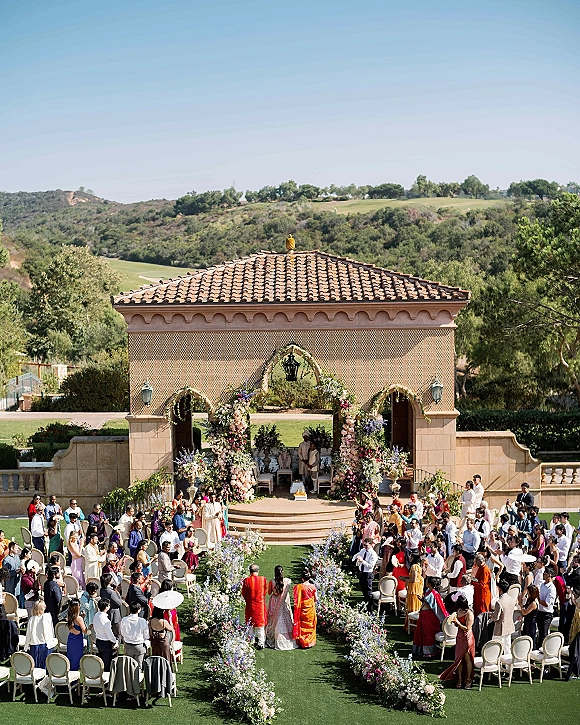 Ceremony setup for an outdoor wedding ceremony with a floral arch and flower-lined aisle on a stone terrace overlooking hills and a stucco villa