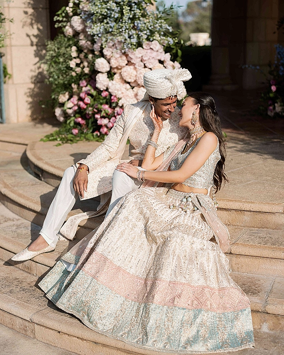 Couple portrait of an Indian wedding couple laughing on stone steps in daylight, bride touching groom’s face in embroidered outfits