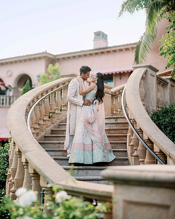 Couple portrait of an Indian wedding couple embracing with a light pink dupatta on a stone villa staircase with arches and palms behind