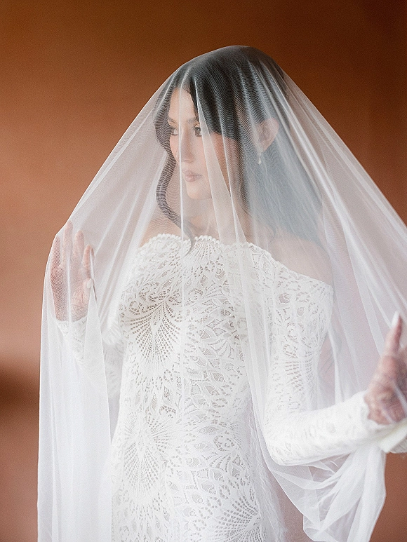 Bridal portrait of a bride under veil, lace wedding dress visible, with drop earrings against a warm brown backdrop