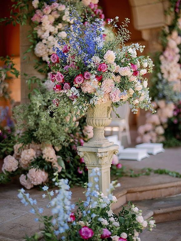 Wedding floral arrangement in a stone urn with garden roses and dahlias, set on stone steps near columns and a floral arch backdrop