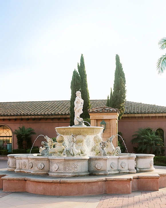 Courtyard fountain at a wedding venue with stone sculpted figures and water jets, set before a stucco villa with cypress trees