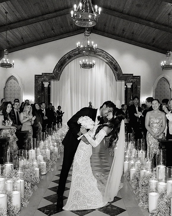 Wedding kiss portrait of bride and groom in a dip kiss, her long veil flowing as guests cheer along a candlelit indoor aisle.