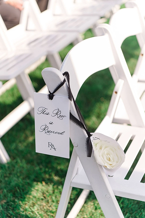 Reserved ceremony seating with a reserved row sign tied in black ribbon and a white rose on white folding chairs along a lawn aisle