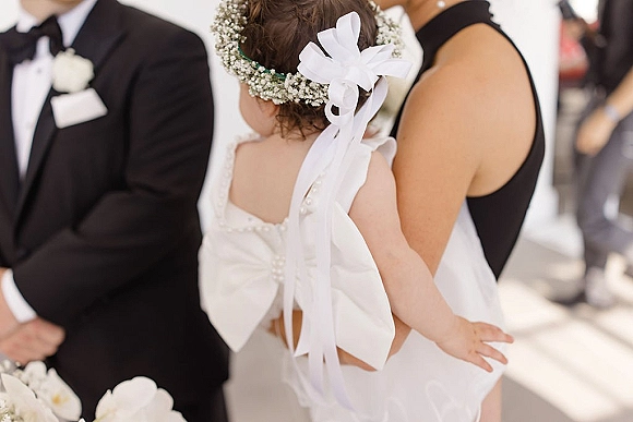 Flower girl in a white dress with a large back bow and baby's breath flower crown, seen from behind at a bright outdoor ceremony