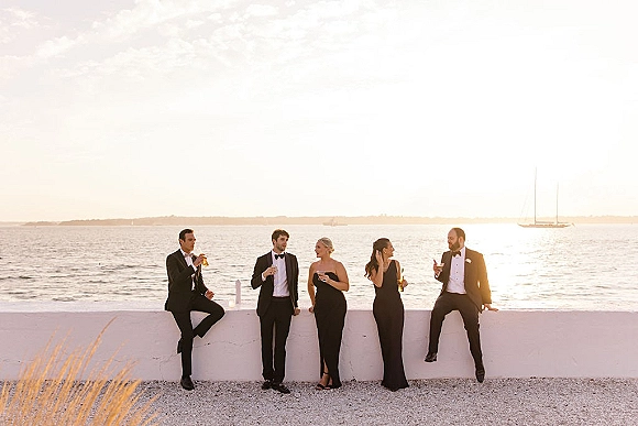 Wedding party portrait of bridesmaids and groomsmen in black tie attire raising champagne glasses by the ocean at sunset with a sailboat