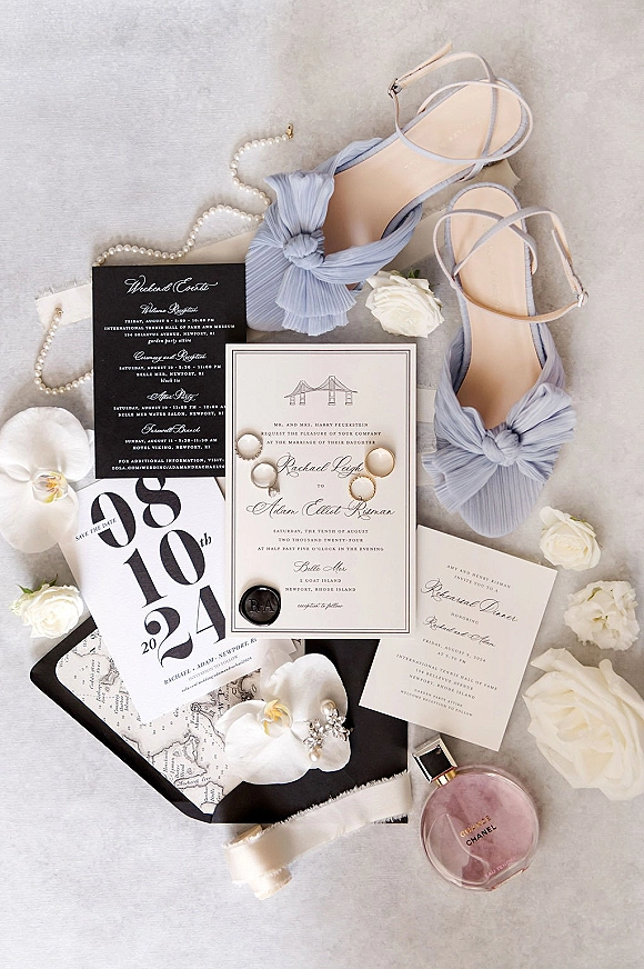 Invitation suite with black and white wedding invitations, wax seal envelopes, pearl jewelry, blue bow heels, and white roses on light fabric