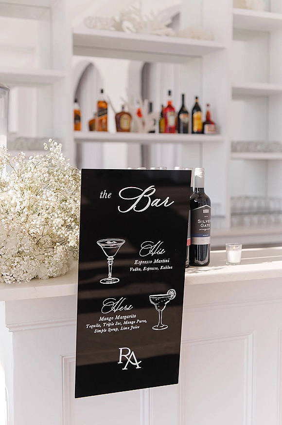 Wedding bar sign with calligraphy cocktail illustration on a white counter, flanked by liquor bottles, votives, and baby's breath blooms