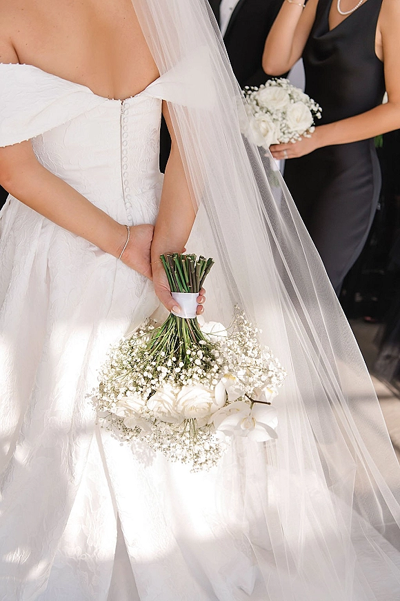 Bridal bouquet with white orchid bouquet and baby’s breath, held behind the bride’s back with long veil and pearl necklace indoors