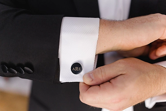 Groom cufflinks with monogram cufflinks on a white dress shirt cuff beneath a black tuxedo sleeve in soft indoor light