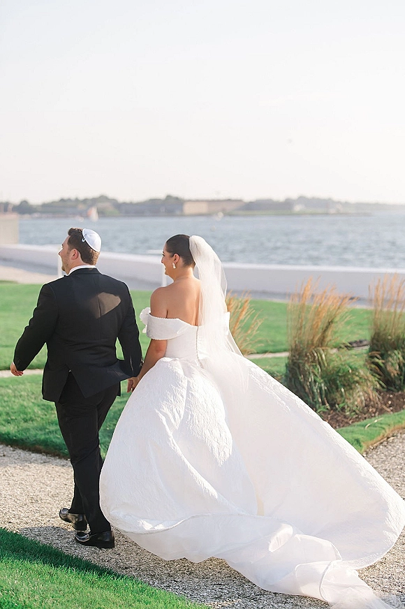 Newlywed couple holding hands, bride and groom walking away in a long train and veil beside a waterfront lawn and seawall
