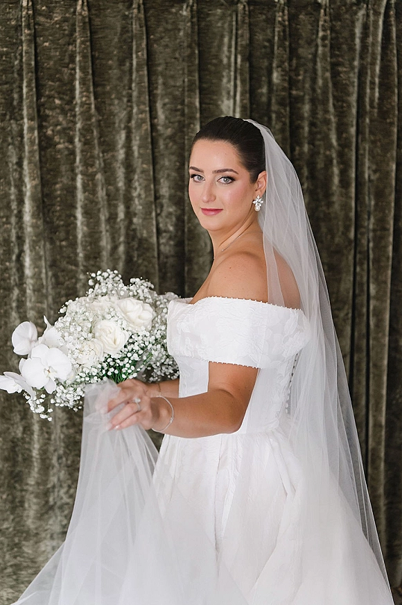 Bridal portrait of a bride in an off the shoulder wedding dress with long veil, holding a white rose bouquet before a velvet curtain backdrop