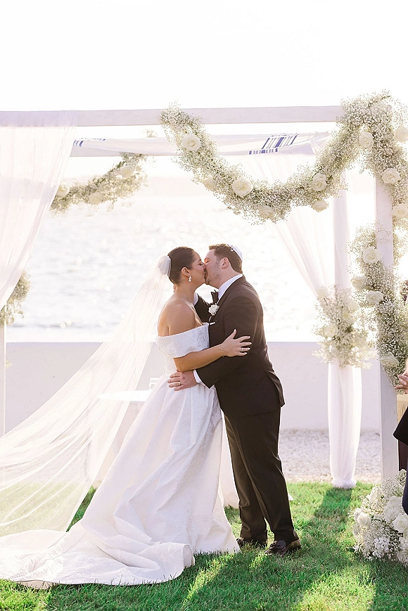 Wedding kiss as bride and groom embrace beneath a white-draped floral arch, veil lifting in ocean breeze on a waterfront lawn