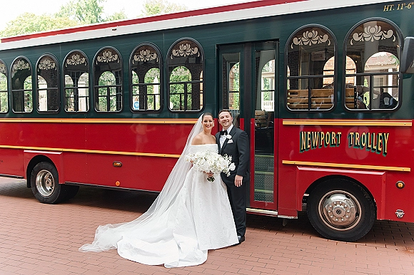 Couple portrait of bride in strapless gown with long veil holding bouquet beside groom in tuxedo by a red trolley on brick pavement