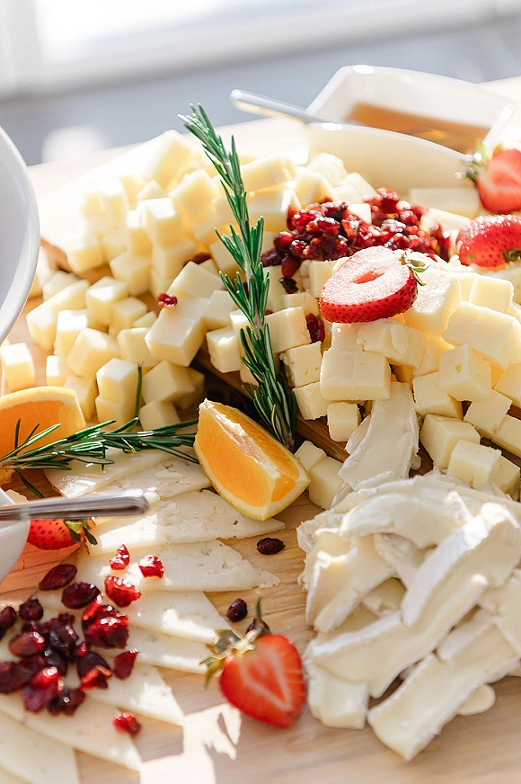 Cheese board with brie, cubes, strawberries, dried cranberries, and a small sauce bowl on a wooden board with white dishware backdrop