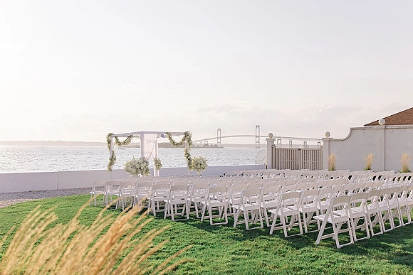 Outdoor ceremony setup with white folding chairs facing a draped arch with floral arrangements and greenery garland by the waterfront lawn