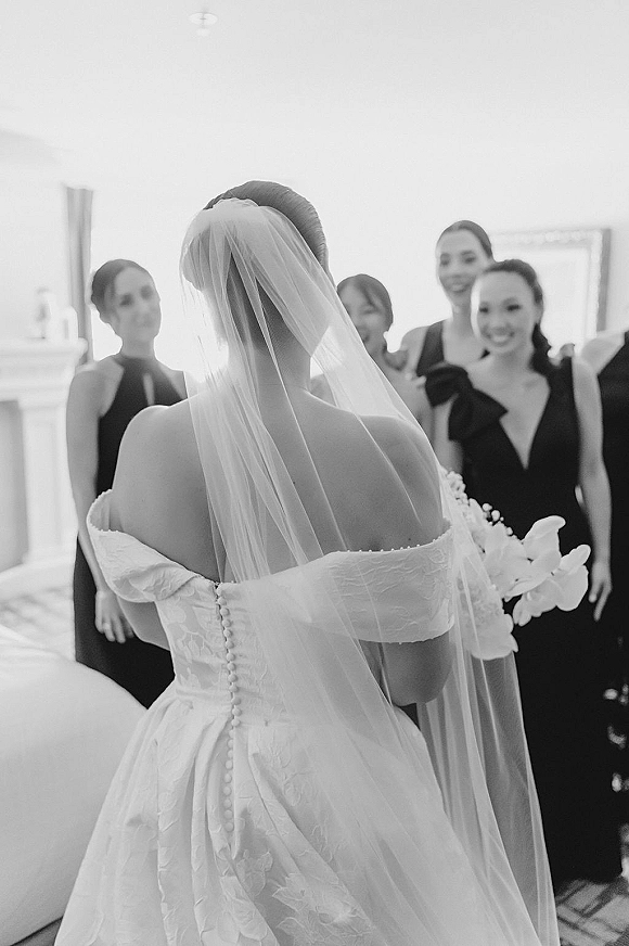 Bride getting ready in a cathedral veil and lace off-the-shoulder gown, holding orchids as bridesmaids react in a hotel room window light