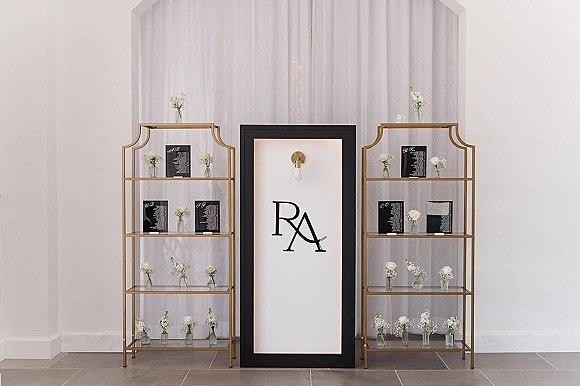 Wedding escort card display on gold shelving with black escort cards, clear acrylic shelves, and white bud vases against white drapery