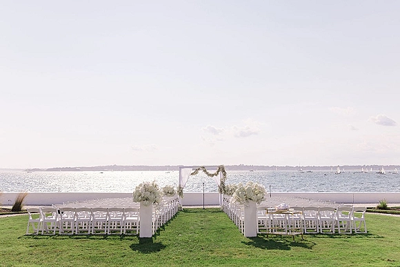 Ceremony setup with white folding chairs lining a grass aisle toward a draped wedding arch, ocean horizon and sailboats beyond