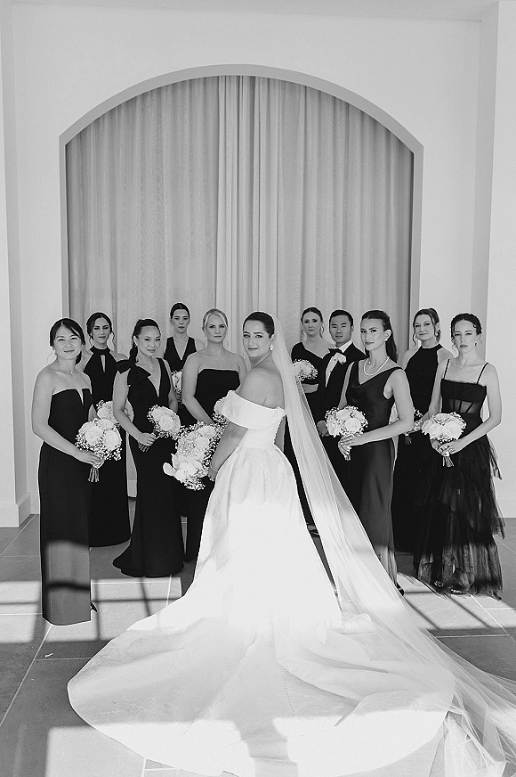 Wedding party portrait with bride and bridesmaids in black dresses, holding white rose bouquets, posed by an arched doorway with draped curtains
