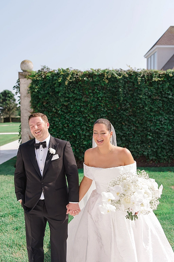 Couple portrait of bride and groom holding hands, laughing as she carries a white orchid bouquet before a green hedge wall in daylight