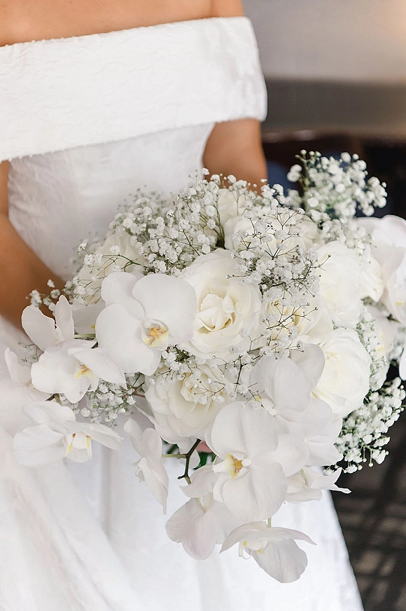 Bridal bouquet of white orchids and roses with baby's breath held against a bridal gown in a softly blurred indoor setting