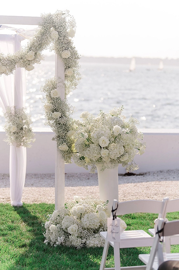Wedding ceremony arch with a white floral ceremony arch of roses and hydrangeas, draped in white fabric on a lawn by the ocean with sailboats