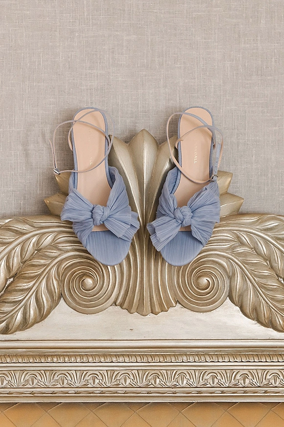 Wedding shoes in dusty blue bridal sandals with bow detail and ankle straps on a carved gilded bench against linen backdrop