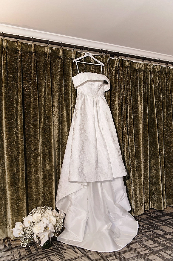 Wedding dress with lace bodice hanging from a rod beside a white orchid and baby’s breath bouquet against velvet drapes indoors
