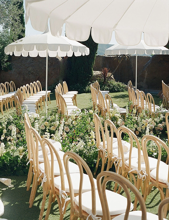 Ceremony setup with wooden chairs and white parasols lining a flower-bordered aisle on a grass lawn beside a stone wall and trees