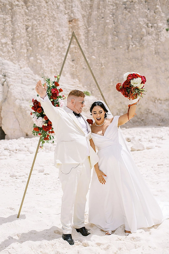 Couple portrait of bride holding bouquet of red and white roses beside groom in a white suit under a geometric arch on white sand cliffs