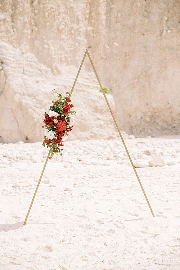 Wedding ceremony arch with a triangle wedding arch frame, red and white flowers and greenery on white sand by a rocky cliff wall