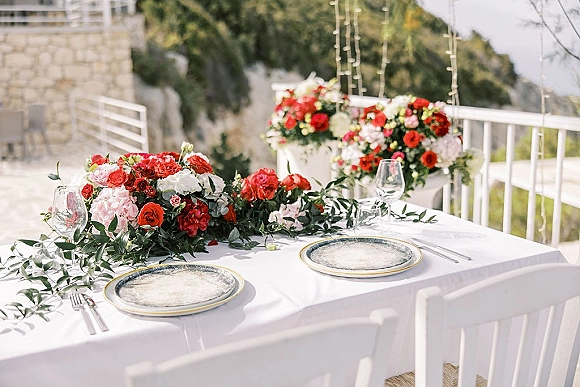 Reception tablescape with head table decor, white linens, red and pink rose centerpiece, place settings and wine glasses on an outdoor terrace