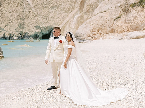 Couple portrait at a beach wedding, bride in veil and off-the-shoulder gown with long train beside groom in white tuxedo by rocky cliffs
