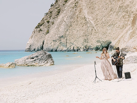 Wedding live music with a wedding singer holding a microphone and acoustic guitar on a beach shoreline with ocean and rocky cliffs behind