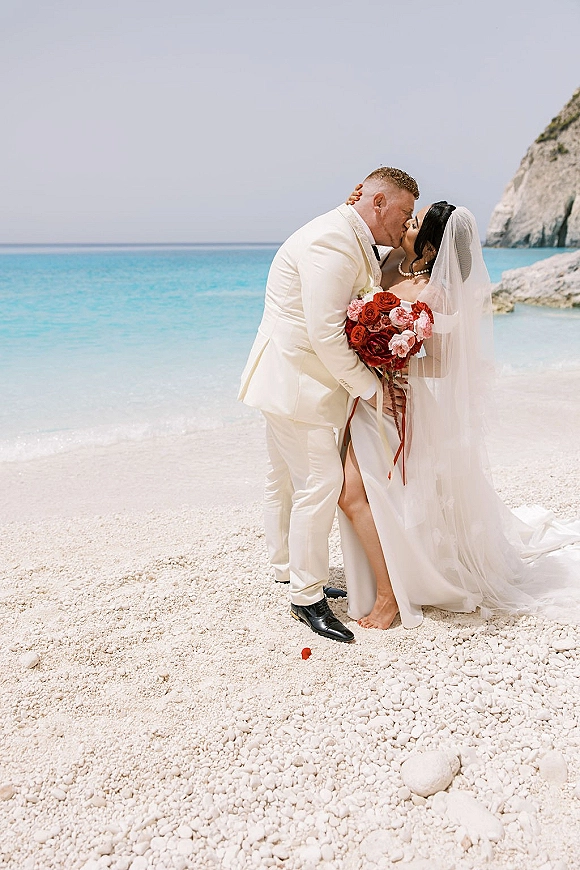 Wedding kiss portrait of bride and groom kissing, bride holding a red bouquet with long veil on a rocky ocean beach cliff backdrop