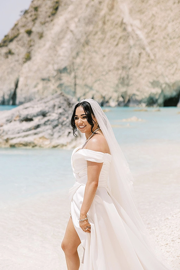 Bridal portrait of a beach bride in a veil, smiling in side profile with pearl necklace on a shoreline by ocean and rocky cliffs
