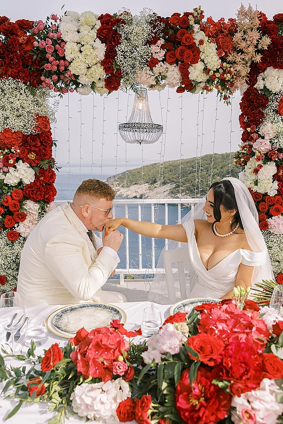Couple portrait of groom kissing bride’s hand at a sweetheart table beneath a floral arch, with chandelier and ocean-view terrace backdrop