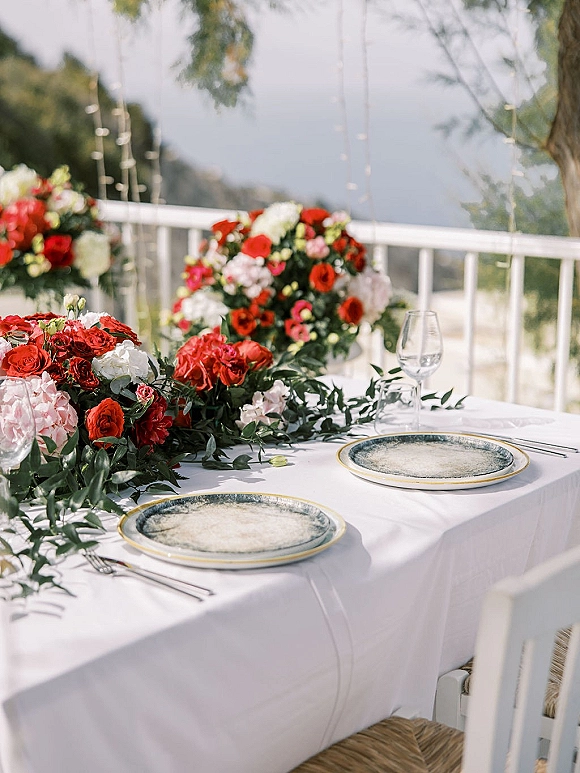 Reception tablescape with a wedding table centerpiece of red and white flowers and greenery garland on a terrace with hills beyond