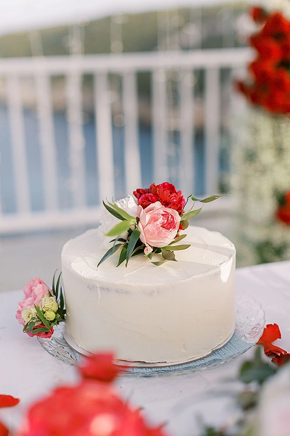 Wedding cake with smooth white buttercream frosting and floral topper of pink peony, red blooms, and greenery by a waterside railing