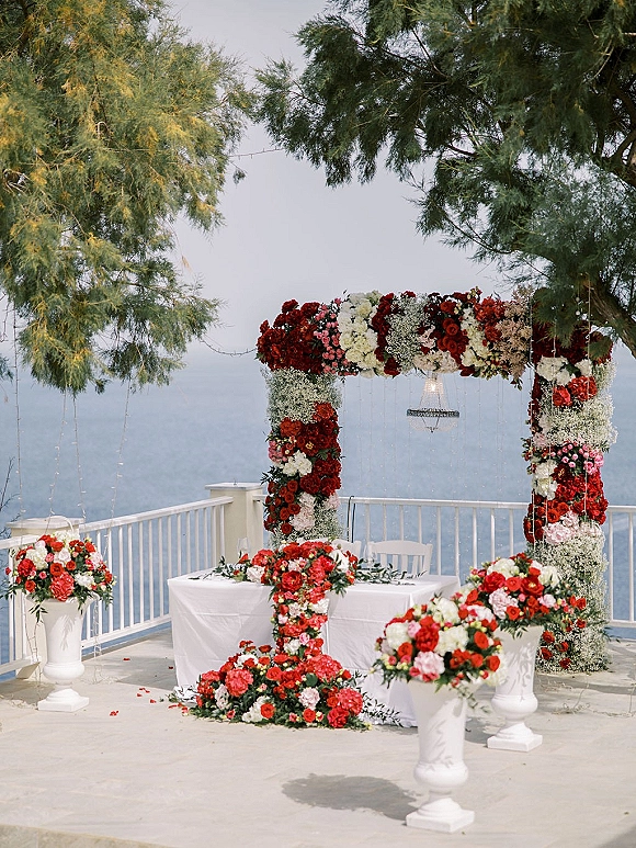 Ceremony setup with wedding floral arch of red and white roses, chandelier and string lights on a terrace patio overlooking the ocean view