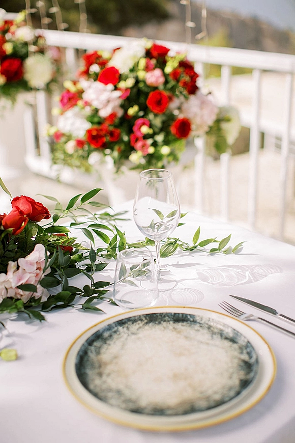 Reception tablescape with wedding table setting on a white tablecloth, greenery garland and red-pink floral centerpiece on an outdoor patio