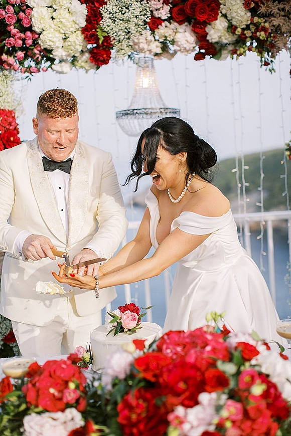 Cake cutting moment as bride and groom laugh by a rose-topped wedding cake under a floral arch with chandelier, ocean view behind