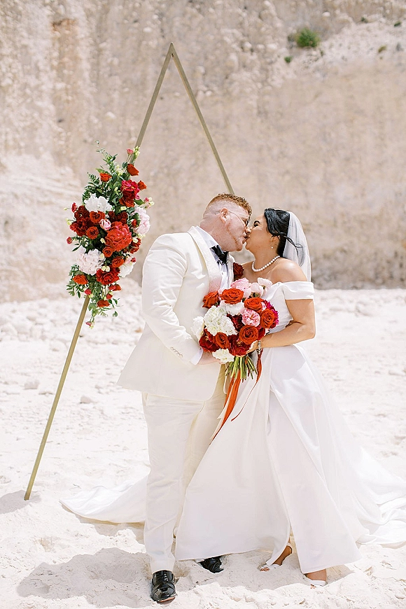 Wedding kiss as bride and groom embrace under a floral triangle arch, veil flowing, holding red and white bouquet on sandy cliffside