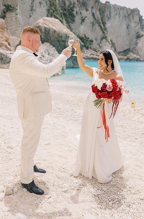Wedding toast as bride and groom clink champagne flutes, bouquet and veil visible on a beach with turquoise water and rocky cliffs