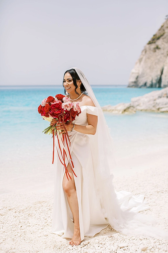 Bridal portrait of a bride holding bouquet in an off-the-shoulder gown and veil on a beach with ocean and rocky cliff behind