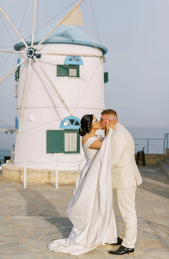 Wedding kiss portrait of bride and groom kissing, bride in off-shoulder gown with long train and sunglasses by a seaside windmill