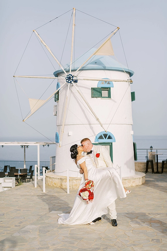 Wedding kiss portrait of groom dipping bride in sunglasses, bouquet in hand, on a stone terrace with windmill and ocean view behind