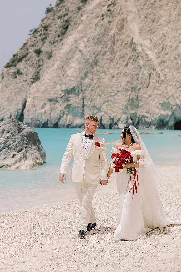 Couple portrait of bride and groom at a beach wedding, holding hands by rocky cliffs and ocean, her red rose bouquet with ribbons and veil flowing