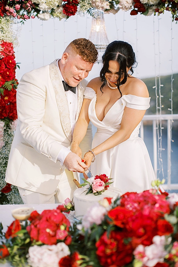 Wedding cake cutting as bride in off-the-shoulder gown and groom in white tuxedo slice buttercream cake beneath chandelier and floral arch